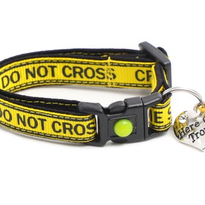 Op de afbeelding: Gele en zwarte hondenriem met de tekst "DO NOT CROSS" en een hartvormig label met de tekst "Here Comes Trouble".