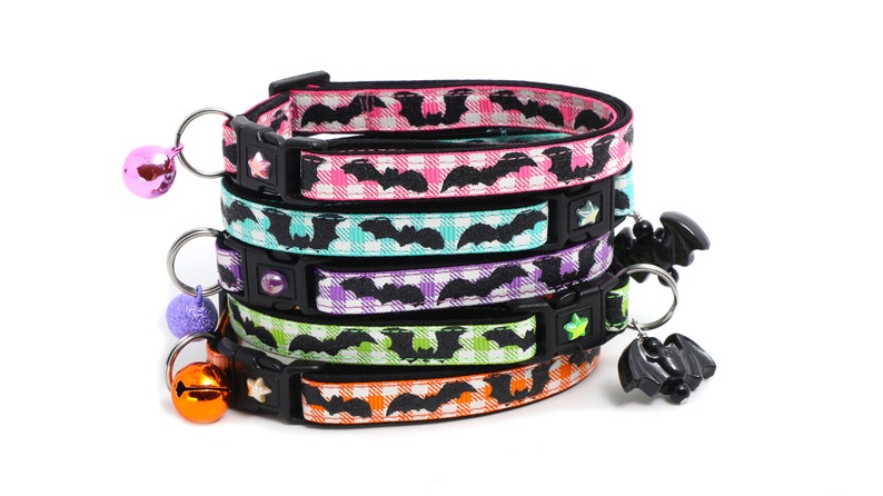 Puede incluir: Una pila de cinco collares para gatos coloridos con un patr&oacute;n de murci&eacute;lagos negros sobre un fondo a cuadros blancos. Cada collar tiene una campana y un amuleto de murci&eacute;lago colgante.
