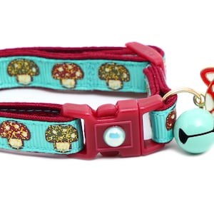 May include: A turquoise blue cat collar with a red mushroom pattern and a red mushroom charm with a white cat face. The collar has a red plastic buckle and a bell.