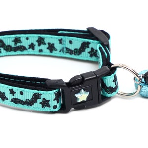 Puede incluir: Un collar para gato azul turquesa con un patrón de murciélago y estrellas negras. El collar tiene una hebilla negra y un anillo plateado con un amuleto de murciélago negro.