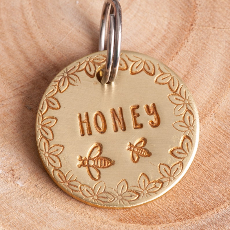 Gold colored round pet tag with darker gold text and design. There are flowers around the edge. The name is straight across the middle. Two Cute bees are under the name.