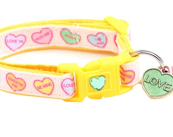 May include: A yellow cat collar with a pink and white ribbon featuring candy hearts with messages like "Love You", "Hug Me", "Sweetie Pie", and "Be Mine". The collar has a yellow plastic buckle and a gold heart charm with the word "LOVE" on it. A pink bell is attached to the charm.