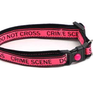 May include: Pink dog collar with black text that reads "Do Not Cross Crime Scene Do Not Cross Crime Scene". The collar has a black buckle and a silver heart-shaped tag that reads "Here Comes Trouble".