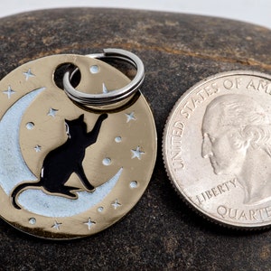 May include: A gold-colored pet tag with a black cat silhouette sitting on a crescent moon. The tag is surrounded by small stars and has a silver ring for attaching to a collar. The tag is next to a US quarter for size comparison.