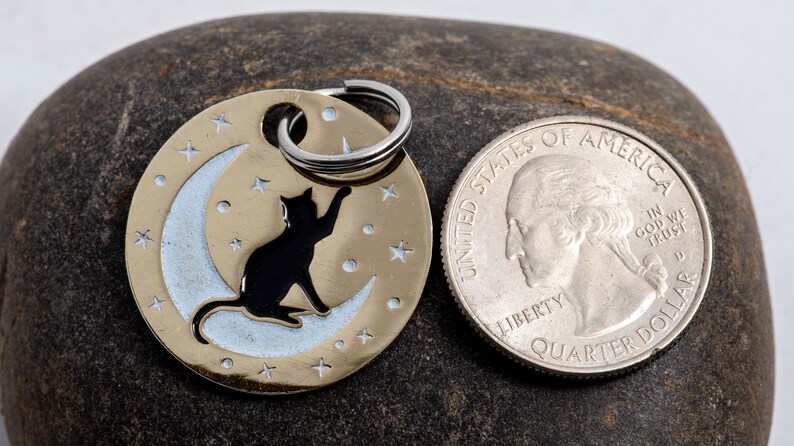 May include: A gold-colored pet tag with a black cat silhouette sitting on a crescent moon. The tag is surrounded by small stars and has a silver ring for attaching to a collar. The tag is next to a US quarter for size comparison.