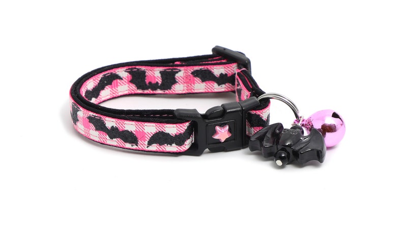 Puede incluir: Collar de gato rosa y negro con un patr&oacute;n de murci&eacute;lago y un amuleto de murci&eacute;lago negro con una campana rosa.