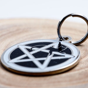 May include: A silver pet tag with a black pentagram and a silhouette of a cat inside. The tag has a silver ring for attaching to a collar.