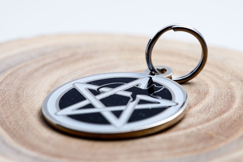 May include: A silver pet tag with a black pentagram and a silhouette of a cat inside. The tag has a silver ring for attaching to a collar.