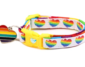 Può includere: Un collare per gatti bianco con un motivo a forma di cuore arcobaleno e una fibbia gialla. Il collare ha un ciondolo a forma di cuore arcobaleno.