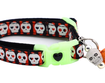 Puede incluir: Collar para gato negro y naranja con un patrón de calavera y huesos cruzados. El collar tiene una hebilla verde y una campana plateada con un dije de calavera.