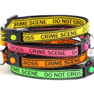 May include: A stack of four colorful cat collars with a black buckle and a bell. The collars are yellow, pink, orange, and green. The collars have the text "Crime Scene Do Not Cross" printed on them. The collars are also decorated with a silver charm that says "Here Comes Trouble".