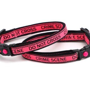 May include: Two pink cat collars with black text that reads "DO NOT CROSS CRIME SCENE". One collar has a pink bell and the other has a silver heart-shaped tag that reads "Here Comes Trouble".