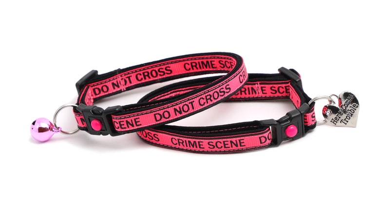 May include: Two pink cat collars with black text that reads "DO NOT CROSS CRIME SCENE". One collar has a pink bell and the other has a silver heart-shaped tag that reads "Here Comes Trouble".