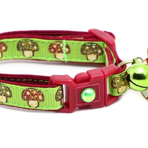 May include: A green and red cat collar with a mushroom print and a red mushroom charm with a cat face. The collar has a red buckle and a green bell.
