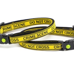 Op de afbeelding: Twee gele en zwarte kattenhalsbanden met de tekst "Crime Scene Do Not Cross" en een zilveren hartvormig label met de tekst "Here Comes Trouble".