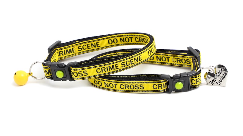 Op de afbeelding: Twee gele en zwarte kattenhalsbanden met de tekst "Crime Scene Do Not Cross" en een zilveren hartvormig label met de tekst "Here Comes Trouble".