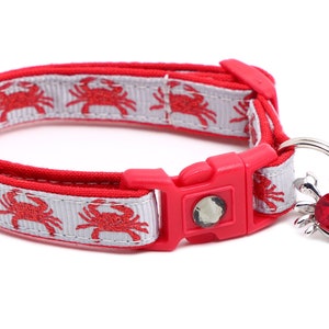 May include: Red and white cat collar with a red crab pattern and a silver charm with a red jewel. The charm is attached to a bell.