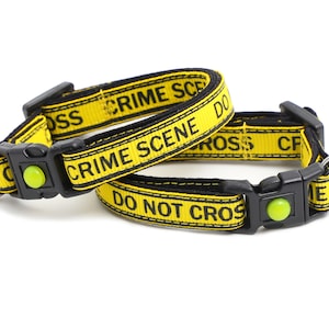 Op de afbeelding: Twee gele en zwarte kattenhalsbanden met de tekst "Crime Scene Do Not Cross" en een zilveren hartvormig label met de tekst "Here Comes Trouble".