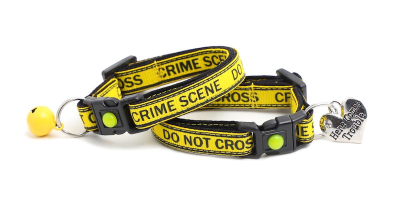 Op de afbeelding: Twee gele en zwarte kattenhalsbanden met de tekst "Crime Scene Do Not Cross" en een zilveren hartvormig label met de tekst "Here Comes Trouble".