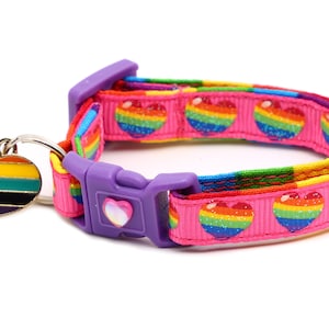 Puede incluir: Un collar para mascotas de color rosa con un patrón de corazón arcoíris y un dije de corazón arcoíris. El collar tiene una hebilla de plástico morado.