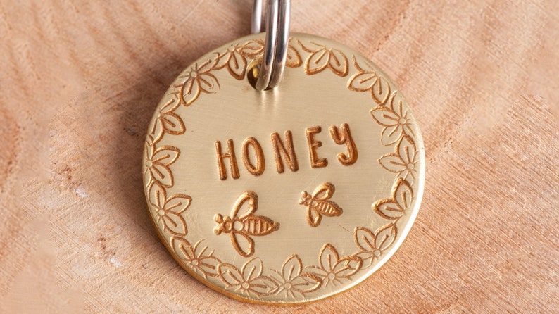 Gold colored round pet tag with darker gold text and design. There are flowers around the edge. The name is straight across the middle. Two Cute bees are under the name.