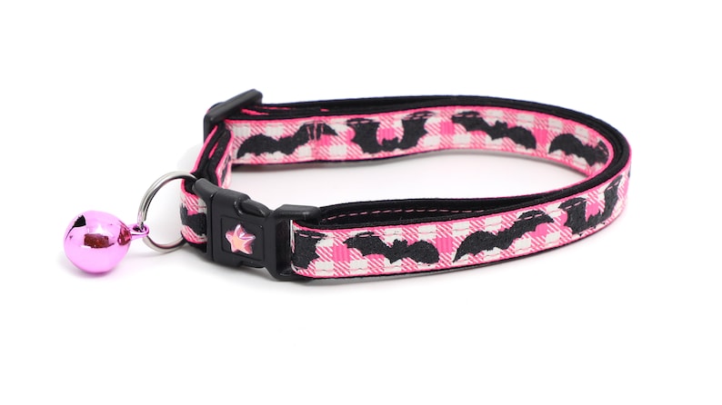 Puede incluir: Collar de gato rosa y negro con un patr&oacute;n de murci&eacute;lagos negros y una campana rosa.
