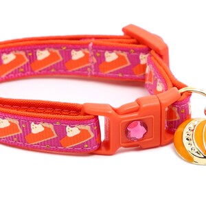 Op de afbeelding: Oranje en roze kattenhalsband met een pompoenbedel en een roze bel. Het halsband heeft een herhalend patroon van taartpunten met een kattengezicht op elke punt.
