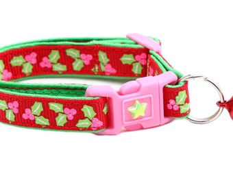 May include: Red and green cat collar with a holly pattern and a pink buckle. The collar has a silver bell attached.