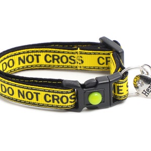 Op de afbeelding: Gele en zwarte hondenriem met de tekst "DO NOT CROSS" erop gedrukt. De riem heeft een zwarte gesp en een zilveren hartvormig label met de tekst "Here Comes Trouble".