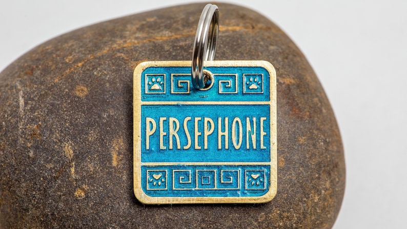 Square pet tag with rounded corners. The tag is gold and light blue. The example name reads Persephone. It is large and in all uppercase letters across the center. Above and below the name is a border of Greek key flourishes and paw prints.