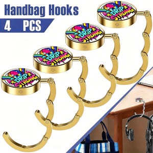 May include: Four gold-colored handbag hooks with a circular top featuring a colorful "DRIP STRONG" design. The hooks are in a folded position, ready to be used. The text "Handbag Hooks" and "4 PCS" are in the upper left corner.