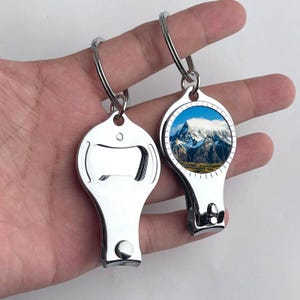 May include: Two silver-toned metal nail clippers with integrated bottle openers and key rings. One clipper features a circular inset graphic of a snow-capped mountain peak under a blue sky, while the other shows the plain polished metal underside.