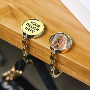 May include: Two gold-colored purse hangers. One has the text "YOUR DESIGN HERE" on a yellow background. The other displays a color photo of a woman. Both have a chain-link design and are attached to a wooden surface.