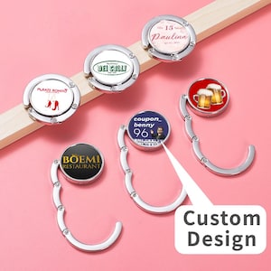 May include: A collection of silver purse hangers with various custom designs. The designs include restaurant logos, text, and images of beer mugs and high heels. The hangers are displayed on a pink surface with a white arrow and the words "Custom Design".