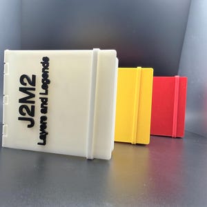 May include: Three rectangular storage cases standing upright in a row. The front case is white with black text reading J2M2 Layers and Legends. The middle case is yellow, and the back case is red. Each case features a vertical strap closure.