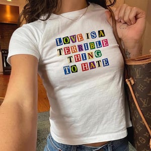 May include: A white short-sleeved t-shirt featuring the text LOVE IS A TERRIBLE THING TO HATE printed in a ransom note style with individual letters in various colors including blue, yellow, red, and green on small square backgrounds.
