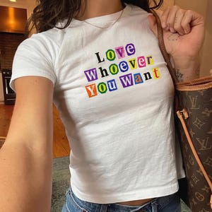 May include: A white short-sleeved t-shirt featuring the text "Love Whoever You Want" printed in a ransom note style with each letter enclosed in a colorful square box. The letters appear in various colors including purple, green, yellow, and orange.