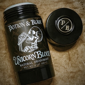 Op de afbeelding: Zwarte deodorantstick met de tekst "POTION & BLADE" en "UNICORN BLOOD". Het etiket toont een schedelillustratie. De deodorant weegt 110 g. Een apart zwart deksel met "PB" is ook zichtbaar.