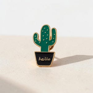 Peut inclure: Une broche en émail et métal doré en forme de cactus vert avec de petits points blancs, planté dans un pot noir. Le mot hello est inscrit en lettres dorées sur le devant du pot noir. La broche présente un contour en métal doré.
