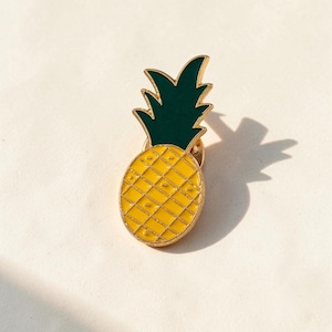 May include: A gold-toned metal enamel pin shaped like a pineapple. The fruit body is bright yellow with a gold grid pattern, and the leafy top is a solid dark green. The pin features a polished gold border and a standard metal clutch backing.