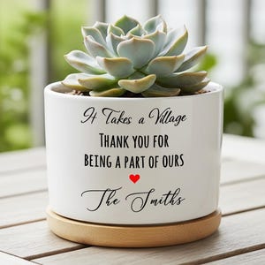 Puede incluir: Una maceta de cerámica blanca con una planta suculenta. La maceta tiene el texto "It Takes a Village, Thank You for Being a Part of Ours" con un corazón rojo y "The Smiths" en escritura. Se asienta sobre una base de madera.
