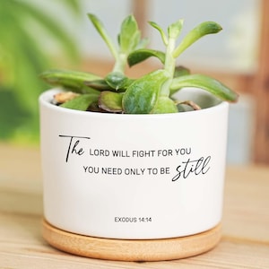 May include: A white ceramic plant pot with a succulent. The pot has the text "The Lord will fight for you, you need only to be still" in black script. It sits on a round wooden base. The text Exodus 14:14 is printed at the bottom.