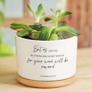 May include: A white ceramic plant pot with a succulent, on a wooden coaster. The pot features the text "But as for you, be strong and do not give up for your work will be reward" and "2 Chronicles 15:7" in black script.