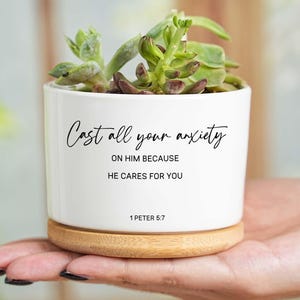 May include: A white ceramic plant pot with a succulent plant, on a wooden base. The pot has the text "Cast all your anxiety on Him because He cares for you" and the Bible verse 1 Peter 5:7.