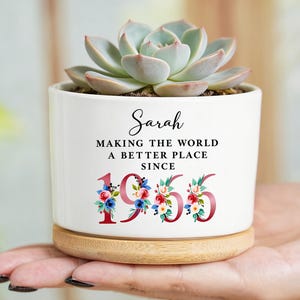 May include: A white ceramic plant pot with a succulent, on a wooden base. The pot reads "Sarah MAKING THE WORLD A BETTER PLACE SINCE 1955" with floral details. The succulent has pale green leaves. The pot is approximately 10 cm in height.