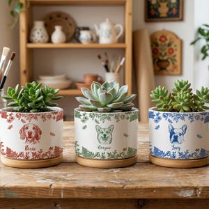 May include: Three ceramic plant pots with succulents, each featuring a different dog breed illustration. The pots are white with red, green, and blue floral designs and dog names. They sit on wooden saucers.