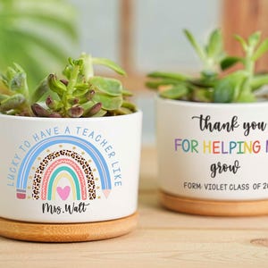 Puede incluir: Dos macetas de cerámica blanca con suculentas. Una maceta tiene un diseño de arcoíris y el texto "Lucky to have a teacher like Mrs. Watt." La otra maceta dice "Thank you for helping me grow, Form Violet Class of 2025."