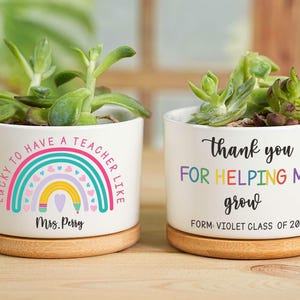 Op de afbeelding: Twee witte keramische plantenpotten met vetplanten. Eén pot heeft een regenboogontwerp en de tekst "Lucky to have a teacher like Mrs. Perry." De andere zegt "Thank you for helping me grow, Violet Class of 2025."