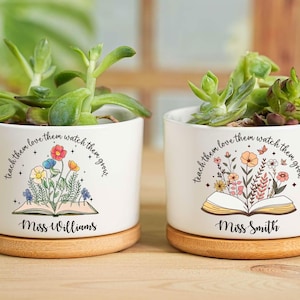 May include: Two white ceramic planters with succulents, each on a wooden base. The planters feature floral designs with an open book and the text "teach them love them watch them grow." One reads "Miss Williams," the other "Miss Smith."