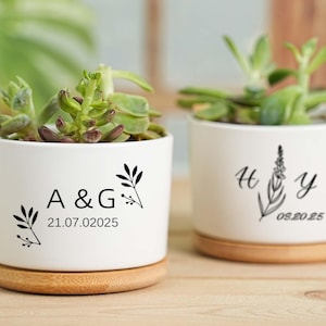 May include: Two white ceramic planters with succulents, each on a wooden base. One planter has "A & G" and "21.07.02025" printed in black, with floral accents. The other has "H Y" and "05.20.25" with a floral design.